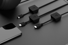 A close-up of a black smartphone lying on a black leather surface next to three different types of black charging cables and adapters. The cables are connected to small, square adapters with a triangular logo.