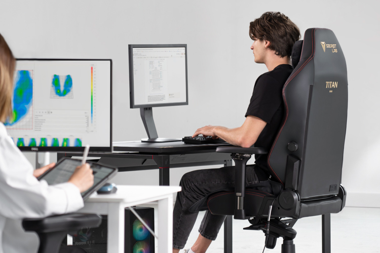 Two people are seated at desks in an office setting. One person, facing away from the camera, is using a computer with a large monitor. The other person, in the foreground, is using a tablet while looking at a second monitor displaying heat maps.