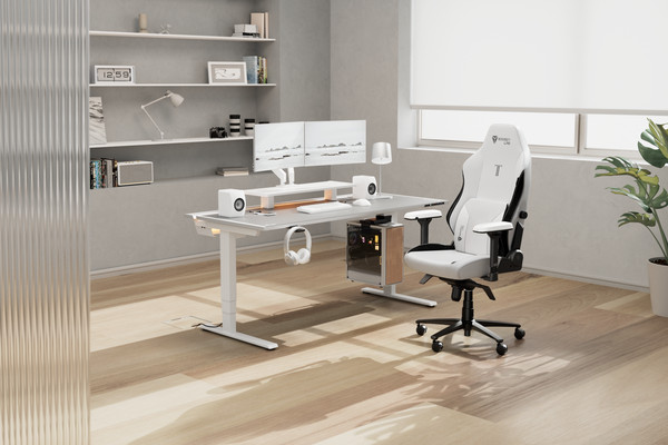 A modern, minimalist home office with a sleek white desk and ergonomic chair. Dual monitors and speakers sit on the desk, alongside a keyboard and mouse. A plant, books, and decor items adorn the surrounding shelves. Natural light streams in from a window.