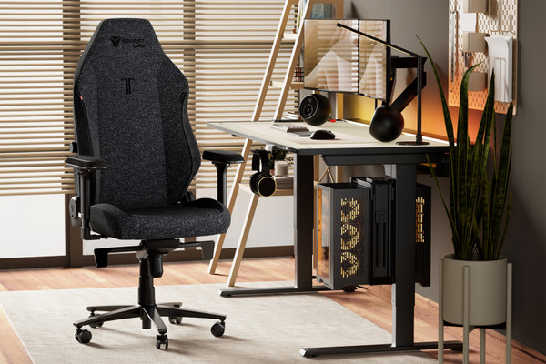 A modern home office setup features a black ergonomic chair with adjustable armrests in front of a desk. The desk holds a computer monitor, keyboard, headphones, desk lamp, and potted plant. A gaming PC sits beneath the desk, and a large plant is placed on the floor nearby.