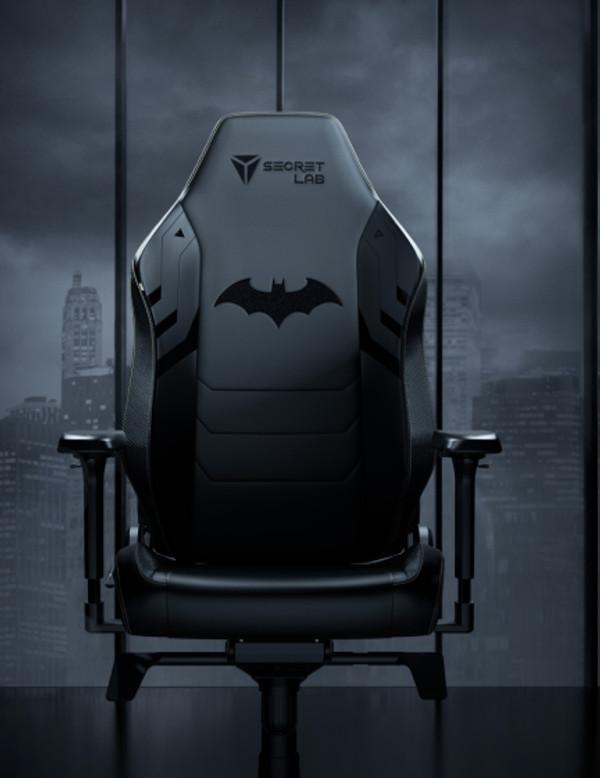 A stylish, black Secretlab gaming chair featuring the Batman logo in the center of the backrest. The chair is situated against a dark cityscape backdrop with tall skyscrapers visible through large windows. The overall color scheme is dark and sleek.