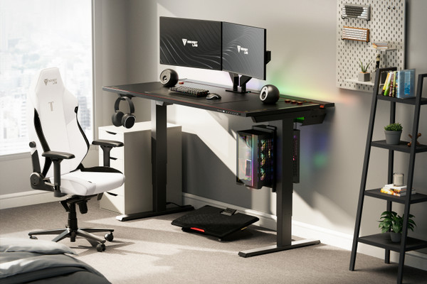A modern gaming setup with a black adjustable standing desk featuring a dual-monitor display, a mechanical keyboard, a mouse, and a gaming chair in white and black. A gaming PC is mounted under the desk. Shelves with books and decor are on the right.