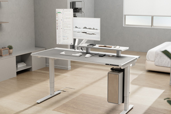 A modern home office setup featuring a motorized standing desk with a sleek design. The desk holds a widescreen monitor on a stand, a laptop, keyboard, mouse, and various gadgets. The background shows a minimalistic room with a bed, shelves, and a window.
